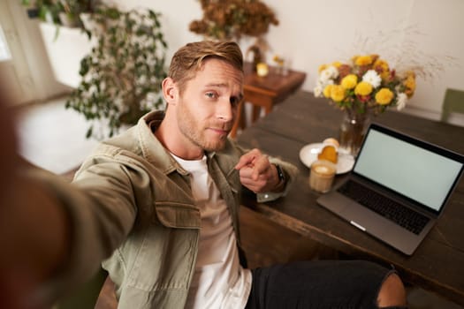 Handsome and confident young man taking selfie on mobile phone, pointing finger at camera, sitting in cafe with laptop, working from coffee shop, looking sassy.