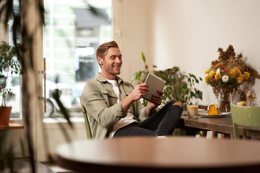 Portrait of happy, relaxed young man sitting in cafe alone, wearing wireless headphones, playing video game on tablet, laughing and smiling, looking at his device. Concept of lifestyle and people.