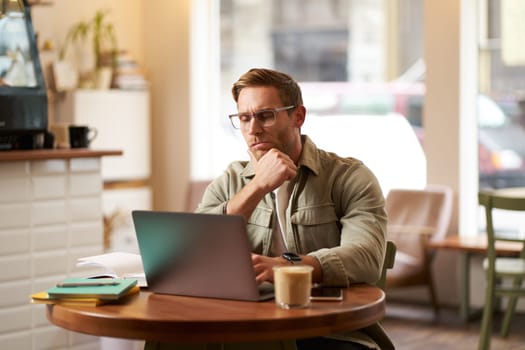 Portrait of businessman in glasses, sits in cafe with laptop, looks concentrated at screen with his project task, works remotely from coffee shop.