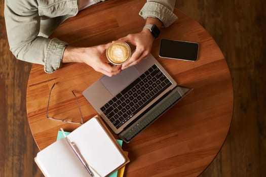 Top view. Male hands holding cup of coffee, man sitting at round table in a cafe, working on laptop, had his smartphone and notebook, drinking cappuccino.