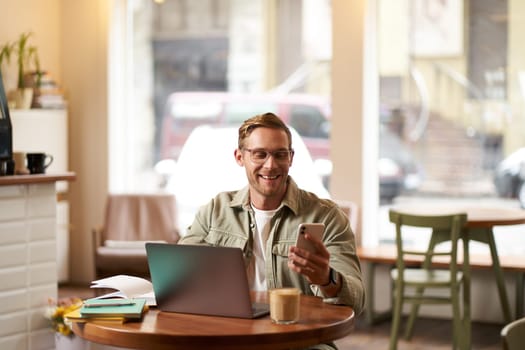 Portrait of handsome man in glasses, sits in cafe with laptop, smiles while reads a message on phone, checks his smartphone app, works from coffee shop.