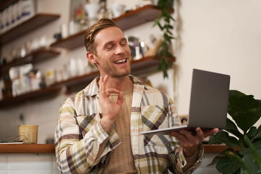Portrait of young smiling man, coworker joins online meeting from cafe, video chats on laptop, shows okay, ok hand sign in approval, drinking coffee and working remotely.