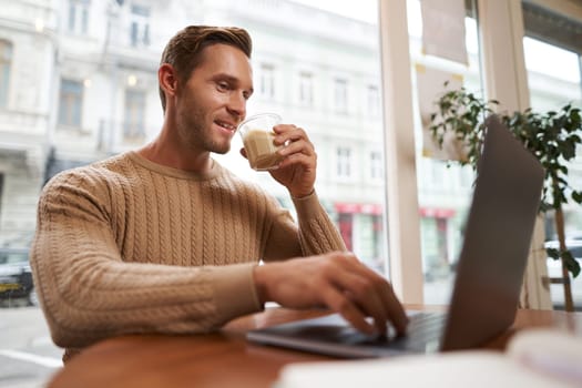 Portrait of young male businessman in cafe, drinking coffee and using his laptop, working from co-working space, sitting near the window and looking at computer screen.