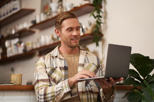 Portrait of young man working online, freelancer sitting in cafe, doing his project on laptop, drinking coffee, smiling while looking at screen.