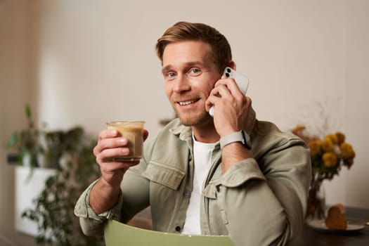 Image of smiling, good-looking young blond man with phone, drinking coffee in cafe, talking to someone over the telephone, answering a call.