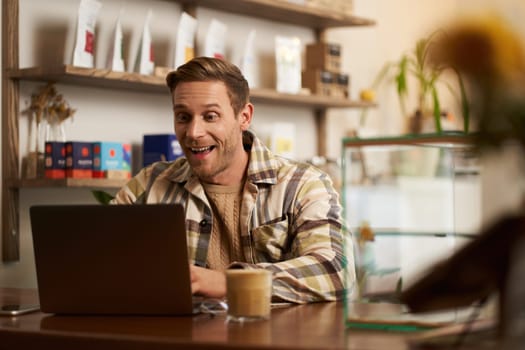 Portrait of handsome young digital nomad, man working in cafe on laptop, looking happy and pleased with his online project, sitting in co-working coffee shop.