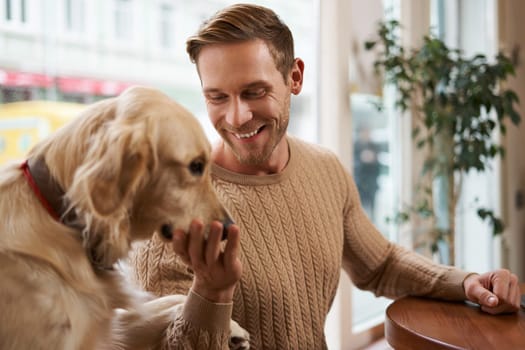 Portrait of handsome smiling blond guy sits in cafe and pets his dog, touches golden retriever.