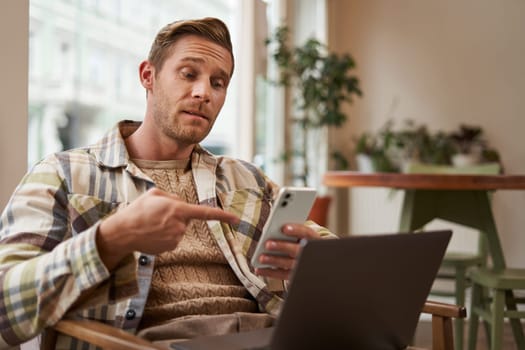 Portrait of young man freelancer, working from cafe, sitting on chair with laptop, pointing finger at his phone screen, showing interest in something on his phone.