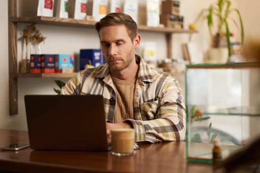 Portrait of young man looking concentrated, working on laptop, freelancer doing his project in cafe, typing on keyboard, drinking coffee in co-working.