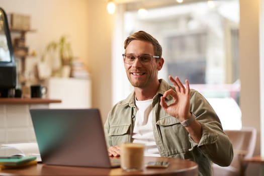 Portrait of handsome businessman in glasses, showing okay hand sign, sitting with laptop in cafe, working remotely, recommends co-working space.