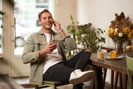 Charismatic, smiling young man, sitting and relaxing in cafe with cup of coffee, listens to music or podcast on wireless earphones, holds smartphone.