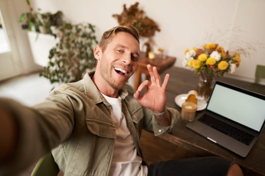 Handsome smiling young man taking selfie, showing okay good hand sign, photographing on mobile phone, sits in cafe with laptop.