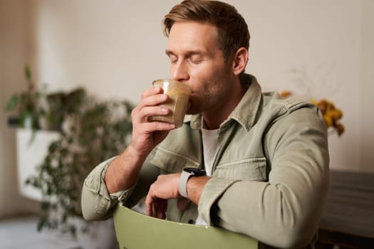 Cafe, leisure and lifestyle concept. Portrait of handsome young man sits on chair with glass of coffee, drinks his beverage and smiling.