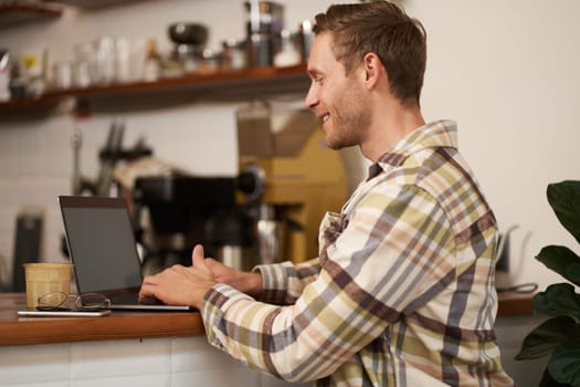 Portrait of handsome smiling man, sitting with laptop in a cafe, typing, working remotely from co-working space, drinking coffee, freelancing from public space.