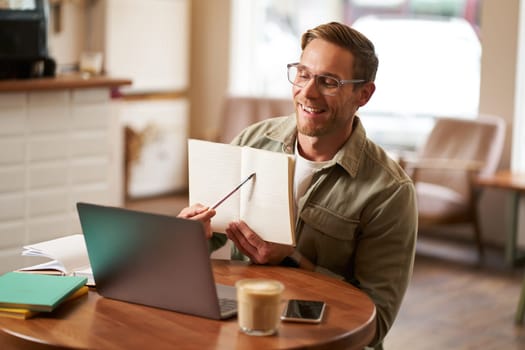 Portrait of handsome man in glasses, working remotely from empty cafe, showing example in his notebook, tutoring, using laptop to meet with student online.