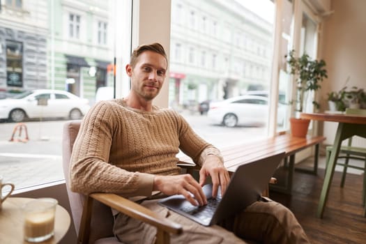 Portrait of young working professional, man sitting in cafe with laptop, drinking coffee and working online, freelancer using coworking space to do his job.