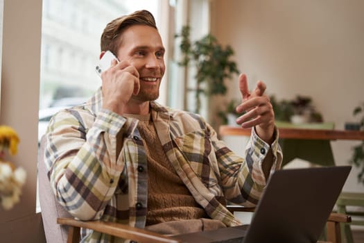 Handsome businessman in coffee shop, sitting in cafe with laptop and calling someone, Man talking on the phone with client, making enquiry about project, talking to coworker while working remotely.