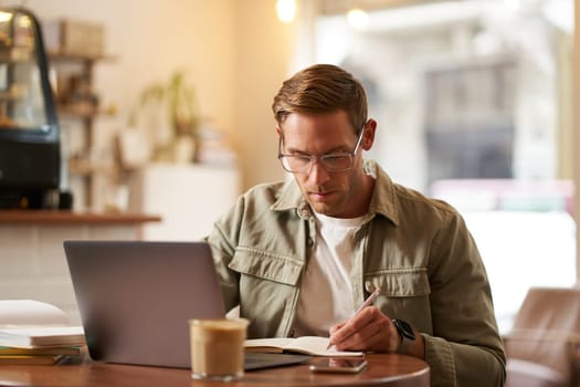Image of focused young man in glasses, sitting in cafe, making notes, studying, attending online course, learning on remote from quite coffee shop.