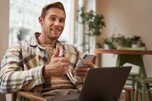 Portrait of smiling, handsome young man relaxing in coffee shop, working remotely from cafe co-working space, using laptop and smartphone, looking at camera and showing thumbs up with satisfied face.