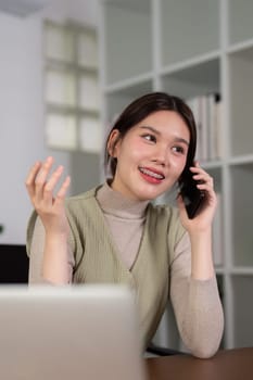 Young woman asian talking on mobile phone while working with laptop at desk home.