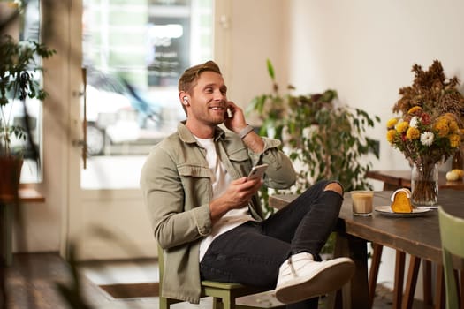 Portrait of young handsome man in casual clothes, sits in cafe and enjoys favourite song, listens to music in wireless headphones, vibing to the relaxing tunes in earphones, holding smartphone.