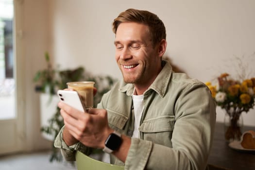 Portrait of smiling charismatic young man, sitting in coffee shop, drinking cappuccino, looking at his mobile phone, reading a message.