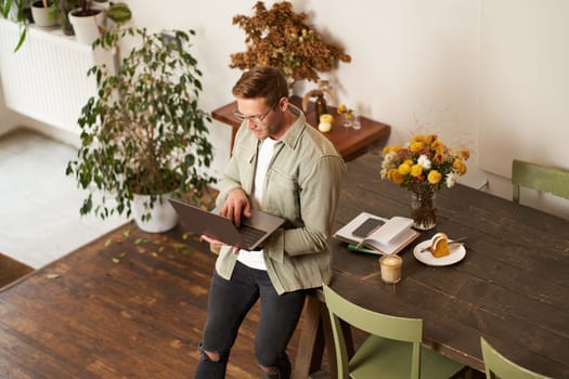 Handsome manager, man in glasses sits on table, holds laptop and types, sends message, works on project, drinks coffee, spends time at co-working space or an office.