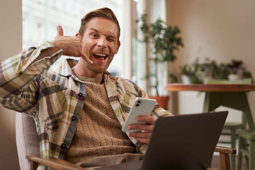 Portrait of handsome male cafe visitor, businessman working on laptop, holding smartphone, showing mobile phone hand sign, give me a call gesture.