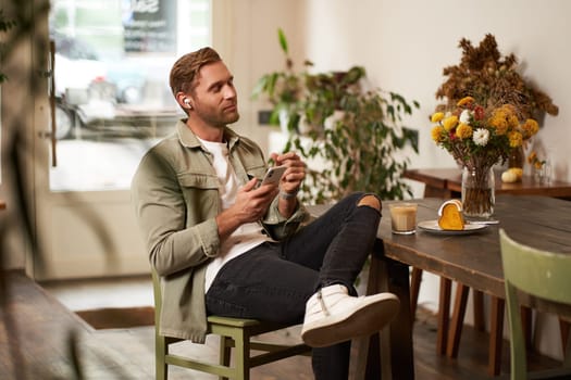 Portrait of young handsome man in casual clothes, sits in cafe and enjoys favourite song, listens to music in wireless headphones, vibing to the relaxing tunes in earphones, holding smartphone.