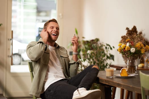 Lifestyle portrait of handsome young man, sitting in cafe, talking to a friend on mobile phone with wireless earphones, laughing and smiling, listening to funny podcast.