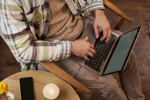 Upper angle cropped shot of male hands, man sitting in cafe with cup of coffee and working on laptop, typing on keyboard, has a phone on the table.