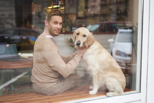 Outdoor shot of a pet-friendly cafe where man sits near the window with his fluffy golden retriever and smiles at camera. Coffee shop visitor with a dog.