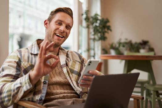 Portrait of handsome man in cafe, sitting with laptop, working remotely, freelancer with phone in hands, shows okay, ok gesture in approval, agree with something, like product.