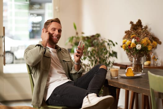 Portrait of handsome, stylish young man, cafe visitor, sitting with his smartphone and listening to music in wireless earphones, smiling at camera.