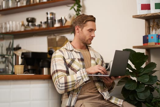 Image of young stylish guy with laptop, sitting in cafe with his computer, typing something, drinking coffee and doing project.