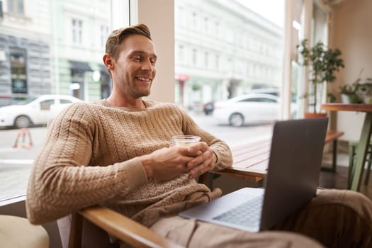 Portrait of handsome young man sits in cafe, drinks coffee and watches video on laptop, looking at screen with happy smile, relaxing in co-working space.