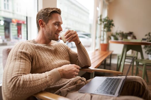 Portrait of handsome modern man, sitting in coffee shop with laptop, looking at screen with happy face, drinking cappuccino, resting in cafe or working.