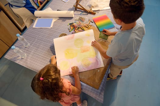 View from above of a teenage school boy and elementary age schoolgirl drawing on paper with pastel color pencils, creating a background for further during art class in the fine art school or gallery