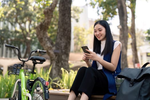 Asian businesswoman with bicycle using smartphone and sitting outside the office building. Woman commuting on bike go to work. Eco friendly vehicle, sustainable lifestyle concept.