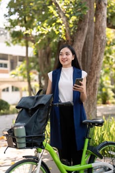 Asian businesswoman with bicycle using smartphone and outside the office building. Woman commuting on bike go to work. Eco friendly vehicle, sustainable lifestyle concept.