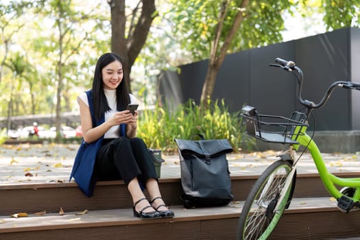 Asian businesswoman with bicycle using smartphone and sitting outside the office building. Woman commuting on bike go to work. Eco friendly vehicle, sustainable lifestyle concept.