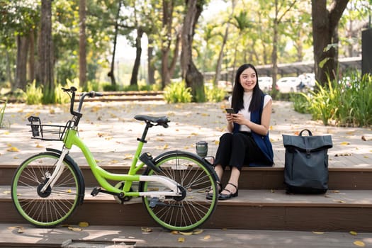 Asian businesswoman with bicycle using smartphone and sitting outside the office building. Woman commuting on bike go to work. Eco friendly vehicle, sustainable lifestyle concept.