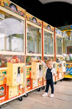 Little girl stands near a claw slot machine filled with toys. High quality photo