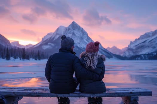 A couple is sitting on a dock overlooking a lake. The man is wearing a black jacket and the woman is wearing a blue jacket. The sky is orange and the mountains in the background are covered in snow