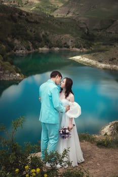 A beautiful wedding couple hugs tenderly against the backdrop of a mountain river and lake, the bride's long white dress.