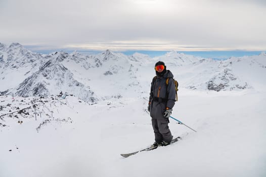 A young man stands on a rock at the top of a mountain with a backpack and skis, watching a long alpine panorama.