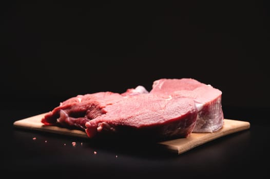 A piece of raw beef in a slice on a wooden cutting board, on a black background. Procuring food.