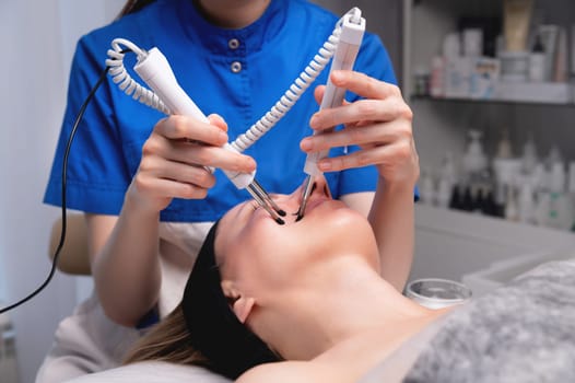 Close-up of a professional cosmetologist's hands touching a woman's cheek with equipment. Young woman lying and relaxing during massage.