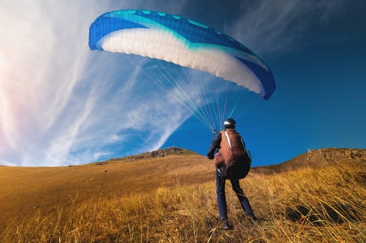 A paraglider stands on yellow grass in a field and holds his parachute in the air. Extreme sport.