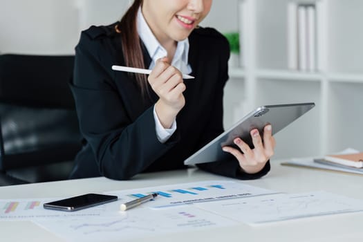 A woman in a business suit is sitting at a desk with a tablet and a pen. She is smiling and she is focused on her work. The scene suggests a professional and productive atmosphere